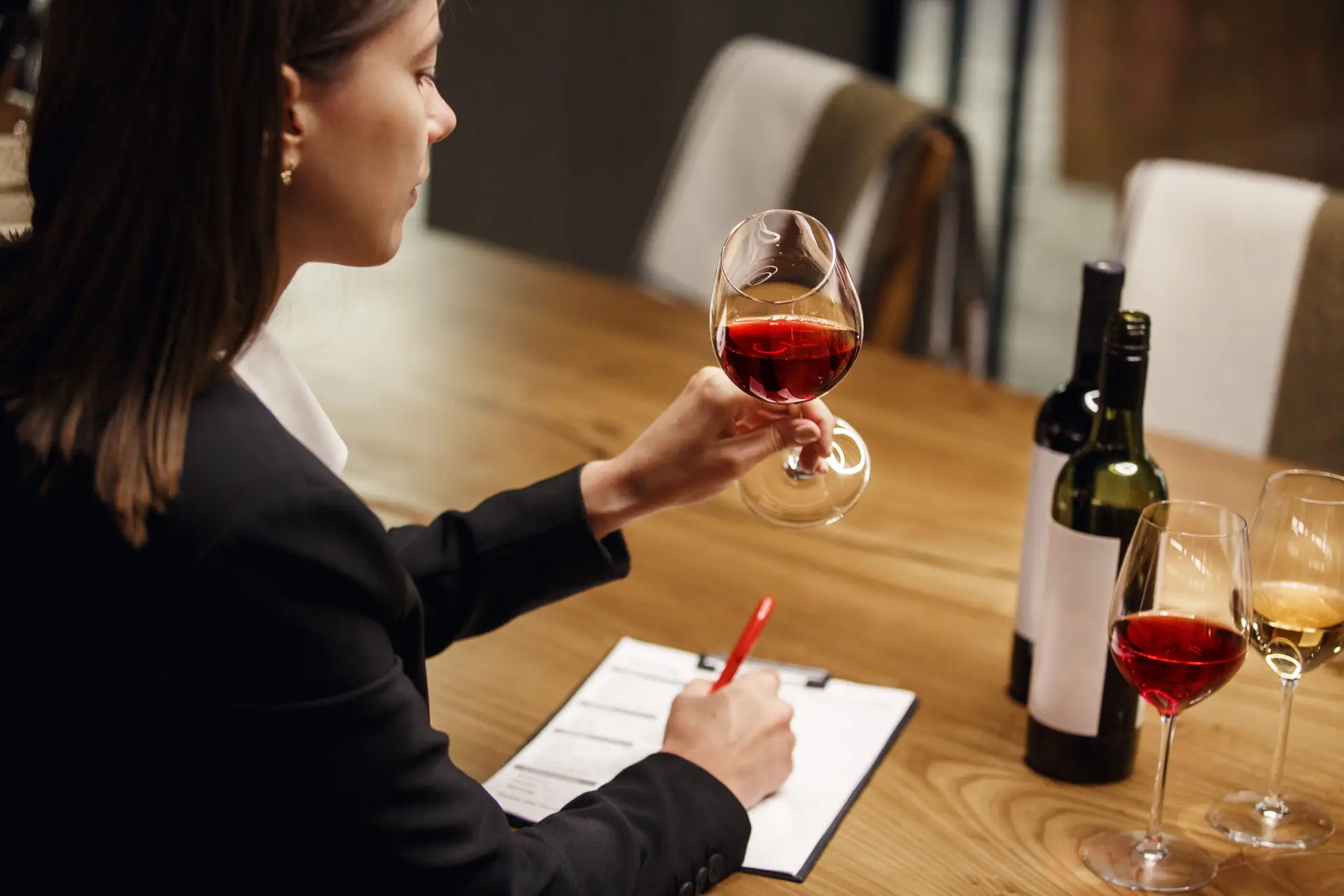 Indoor portrait of young woman wine expert studies red wine looking on the his color sitting at the table being in cellar. Female sommelier tasting red wine during wine courses exam.